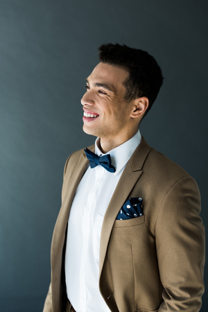 Stylish mixed race man in suit and bow tie smiling isolated on grey backgroundの写真素材