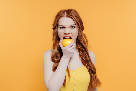 Redhead girl looking at camera, making facial expression and biting lemon isolated on yellow backgroundの写真素材