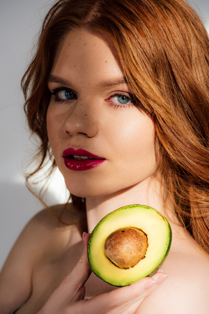 Beautiful redhead girl with red lips looking at camera and posing with cut avocadoの写真素材