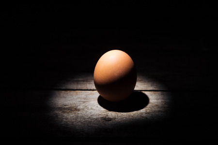 brown chicken egg in darkness on weathered wooden surfaceの写真素材