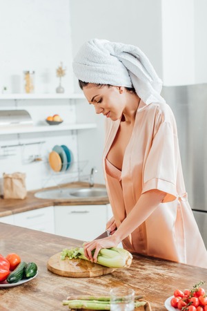 Sexy woman in housecoat with towel on head cutting celery with knife in kitchenの写真素材