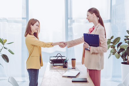 Happy recruiter holding clipboard and shaking hands with woman in modern officeの写真素材