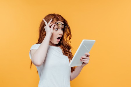 Surprised redhead girl with glasses using digital tablet isolated on yellow background