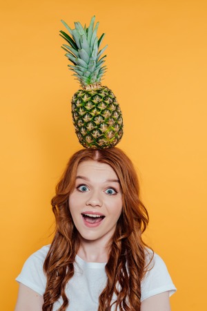 Excited redhead girl looking at camera and posing with pineapple on head isolated on yellow backgroundの写真素材