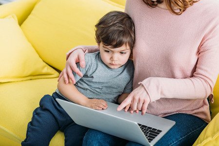 Cropped view of woman with adorable son using laptop while sitting on yellow sofa at homeの写真素材