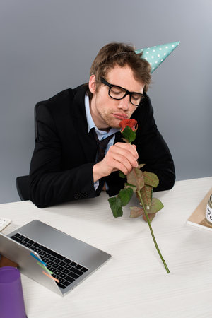 drunk businessman in birthday cap smelling rose at workplace on grey backgroundの写真素材
