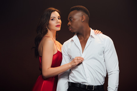Beautiful woman in red dress undressing African American man isolated on black backgroundの写真素材