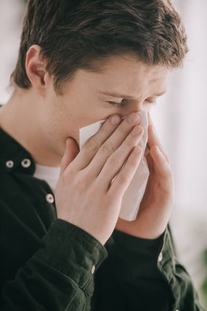 Close up of man having allergy and sneezing in tissue with closed eyes at homeの写真素材
