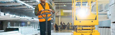 Panoramic shot of Indian worker in safety vest near pallet jack in warehouse