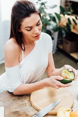 High angle view of pretty stylish girl holding cut avocado near table in kitchenの写真素材
