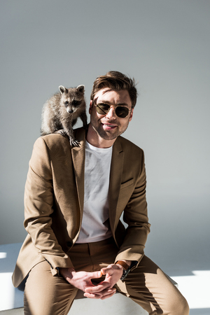 Handsome man in sunglasses with cute raccoon on shoulder on grey backgroundの写真素材