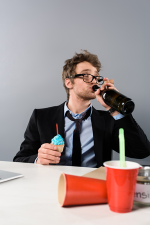 Drunk businessman sitting at workplace and drinking champagne from bottle isolated on grey backgroundの写真素材