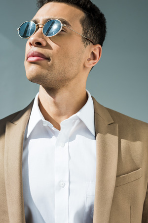 stylish mixed race man in suit and sunglasses isolated on grey with sunlightの写真素材
