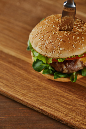 knife in delicious meat burger with cheese, greenery and tomatoes and sesameの写真素材