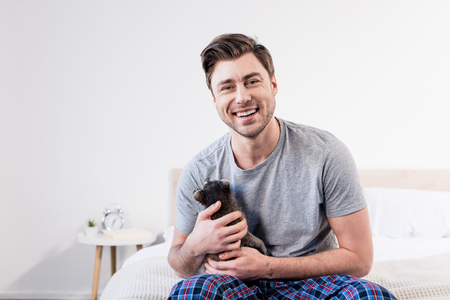 cheerful man in pyjamas sitting on bedding and cuddling with cute raccoonの写真素材