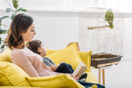 attractive woman with book and cute boy resting on sofa near bird cageの写真素材