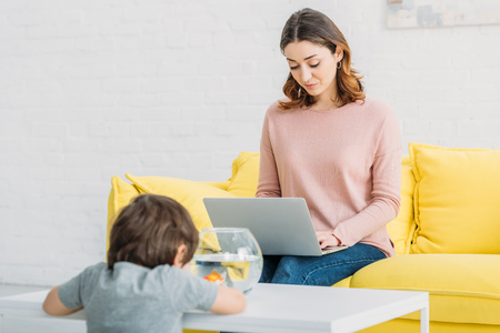 pretty woman using laptop while adorable son looking at fish bowlの写真素材