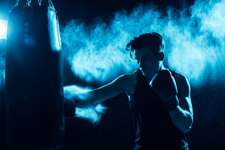 Concentrated boxer in boxing gloves training with punching bag in darkの写真素材