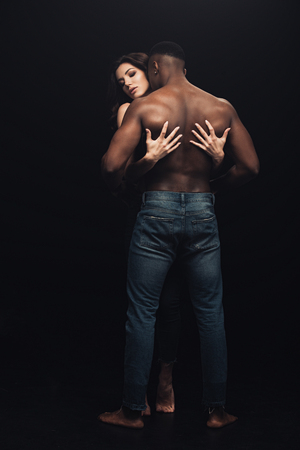 back view of beautiful sexy woman hugging shirtless african american man isolated on blackの写真素材