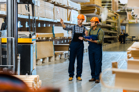 serious storehouse worker in uniform pointing with finger while standing near indian colleague