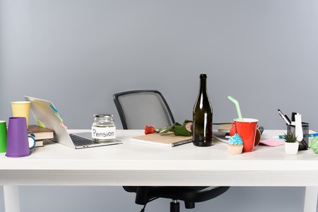 messy workplace with champagne bottle, paper cups, pension moneybox and laptop on white tableの写真素材