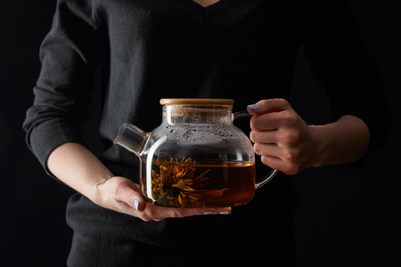 cropped view of young woman holding transparent teapot with chinese blooming tea isolated on blackの写真素材