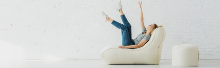 panoramic shot of cheerful blonde girl lying on  bean bag chair and gesturing near brick wallの写真素材