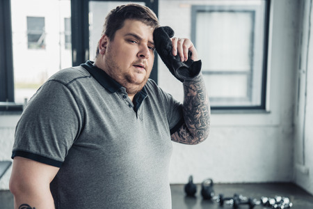tired overweight tattooed man looking at camera and wiping face with towel at sports centerの写真素材