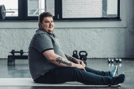 overweight tattooed man sitting on fitness mat and looking at camera at sports centerの写真素材