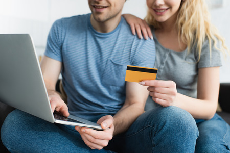 cropped view of blonde woman holding credit card near cheerful man using laptop