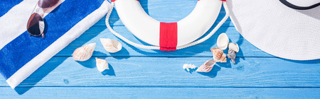 panoramic shot of striped towel, sunglasses, lifebuoy, white floppy hat and seashells on blue wooden...