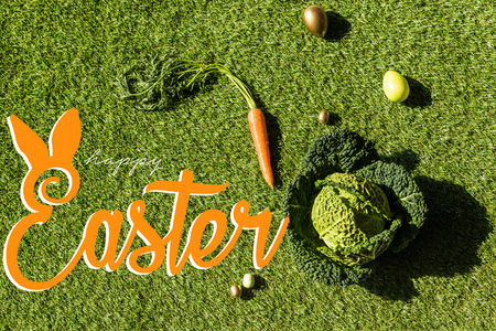 top view of carrot, savoy cabbage and painted Easter eggs on green grass with happy Easter letteringの写真素材
