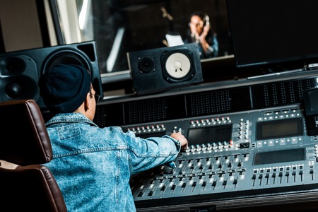 selective focus of mixed race sound producer working at mixing console while pretty woman singing in recording studioの写真素材