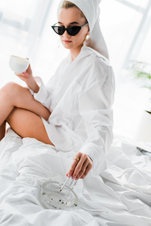 selective focus of young stylish woman in jewelry and sunglasses with towel on head and cup of coffee smoking cigarette with ashtray on bedの写真素材