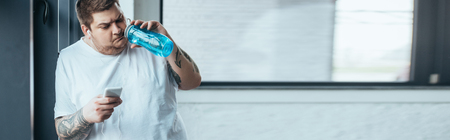 panoramic shot of overweight tattooed man using smartphone and drinking water from sport bottle at gymの写真素材
