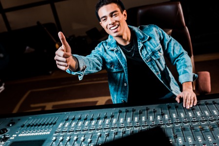 smiling mixed race sound producer showing thumb up while standing near mixing console in recording studioの写真素材
