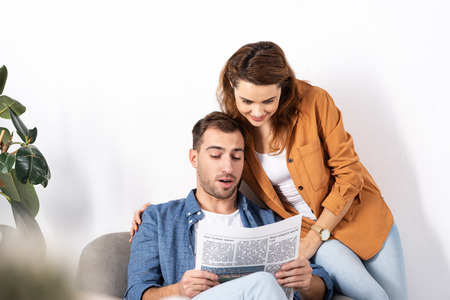 cheerful woman hugging man and reading newspaper at homeの写真素材