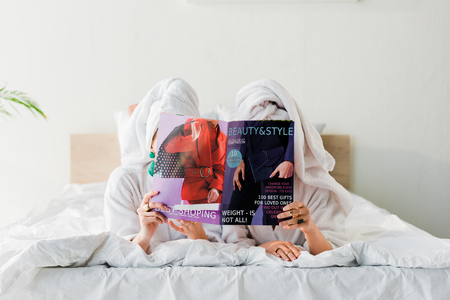 women in bathrobes and jewelry, with towels on heads hiding behind magazine together while lying in bedの写真素材