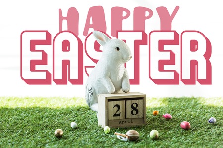 decorative rabbit, wooden calendar with 28 April date, and colorful Easter eggs on green grass with...