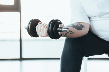 partial view of Overweight tattooed man sitting and training with dumbbell at gymの写真素材