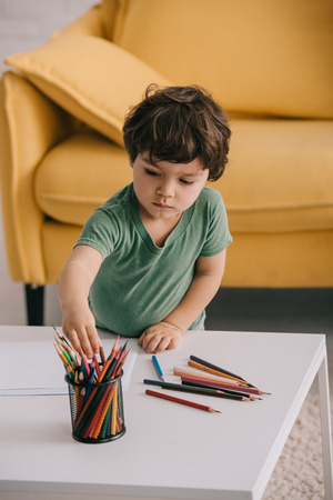 kid in green t-shirt with color pencils and papers in living roomの写真素材