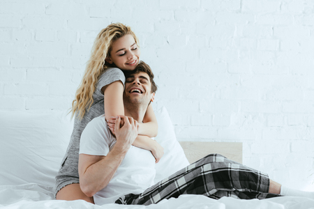attractive and happy blonde girl hugging cheerful boyfriend sitting on bedの写真素材