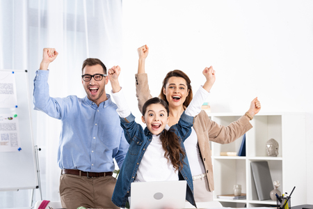 cheerful family celebrating triumph near laptop in officeの写真素材