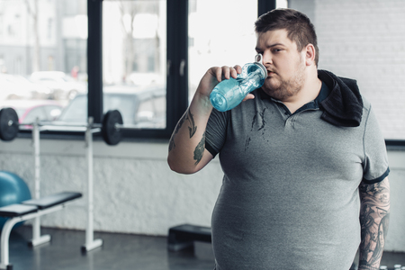 overweight tattooed man with towel drinking water from sport bottle at gymの写真素材