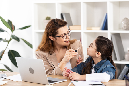 cheerful mother applying lip gloss on cute daughter in officeの写真素材