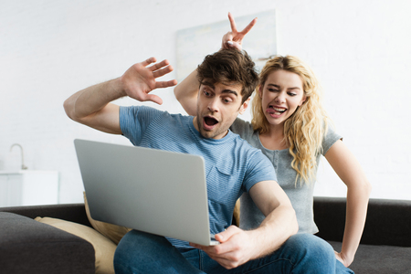 cheerful man waving hand near happy woman showing tongue and peace sign while having video call on laptop