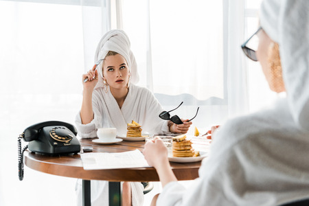 Dissatisfied stylish woman in bathrobe and jewelry with towel on head and sunglasses rolling her eyes during breakfast with friendの写真素材