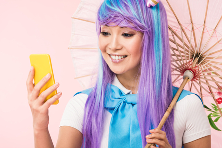 Smiling Asian anime girl with paper umbrella looking at smartphone isolated on pink backgroundの写真素材
