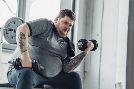 Obese tattooed man exercising with dumbbells and looking at camera at sports centerの写真素材