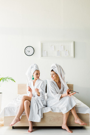 stylish smiling women in bathrobes and jewelry, with towels on heads sitting on bed with nail filesの写真素材